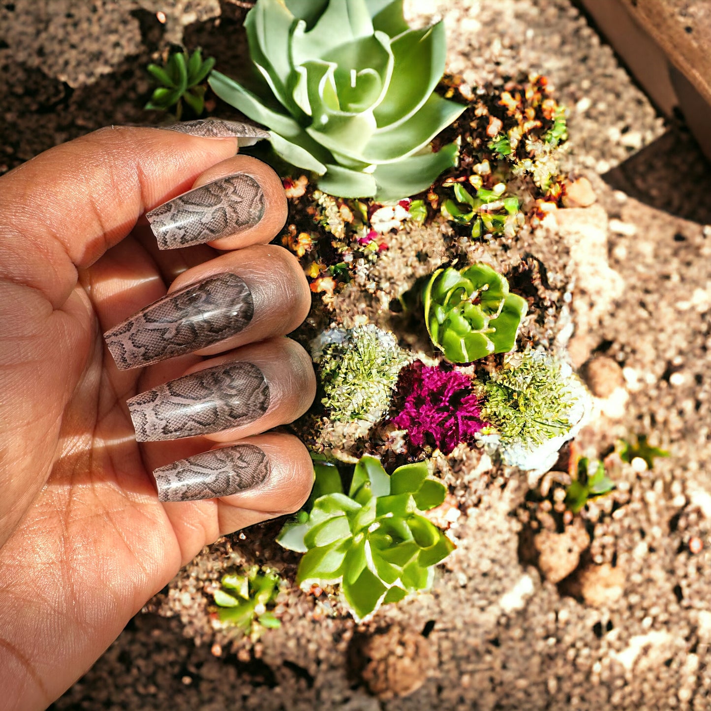 Hand adorned with long coffin-shaped press-on nails featuring a snake skin design against a vibrant flower garden background