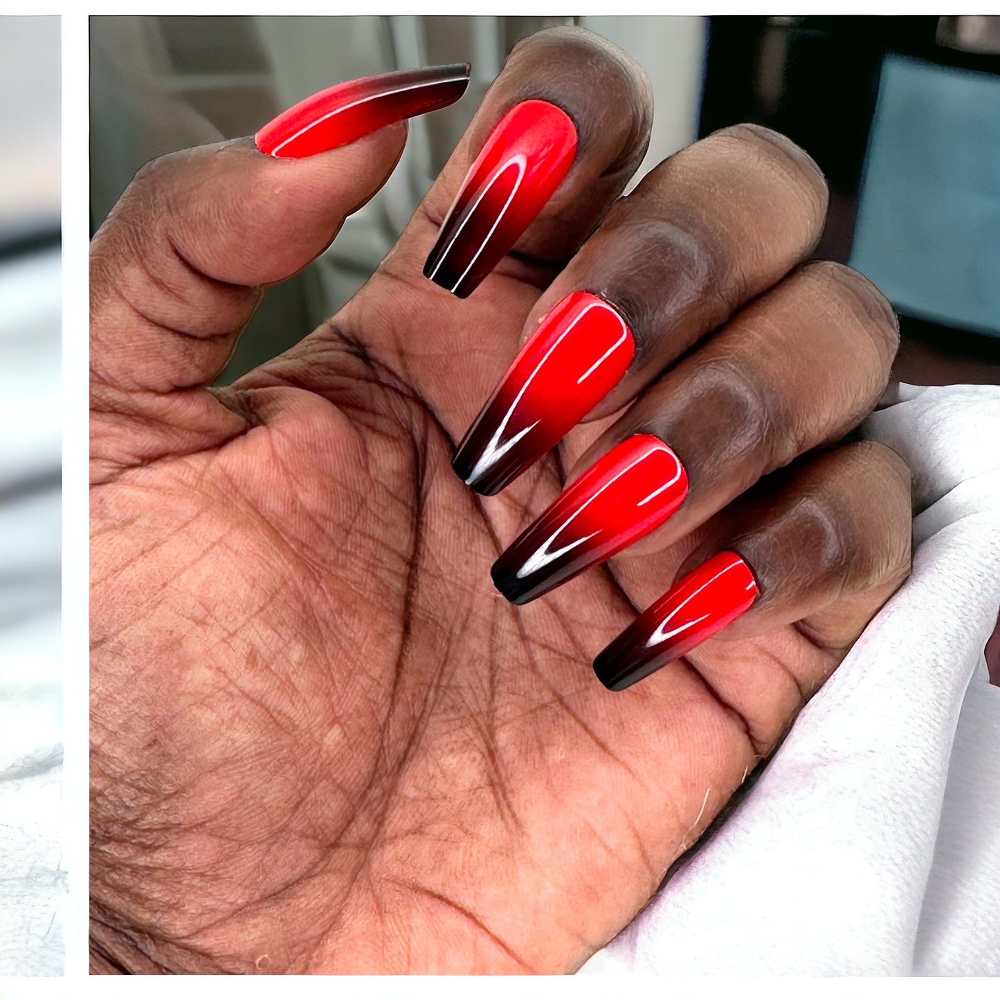 A hand featuring long coffin-shaped press-on nails in a striking red and black design, resting on a white fabric