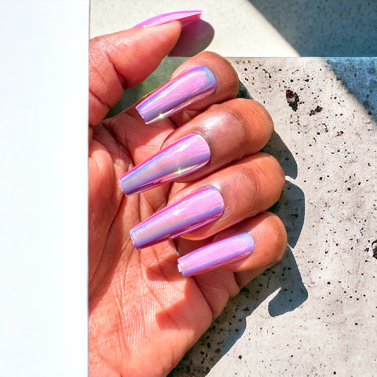 A hand showcasing long coffin-shaped press-on nails in a shimmering pink chrome color against a neutral background