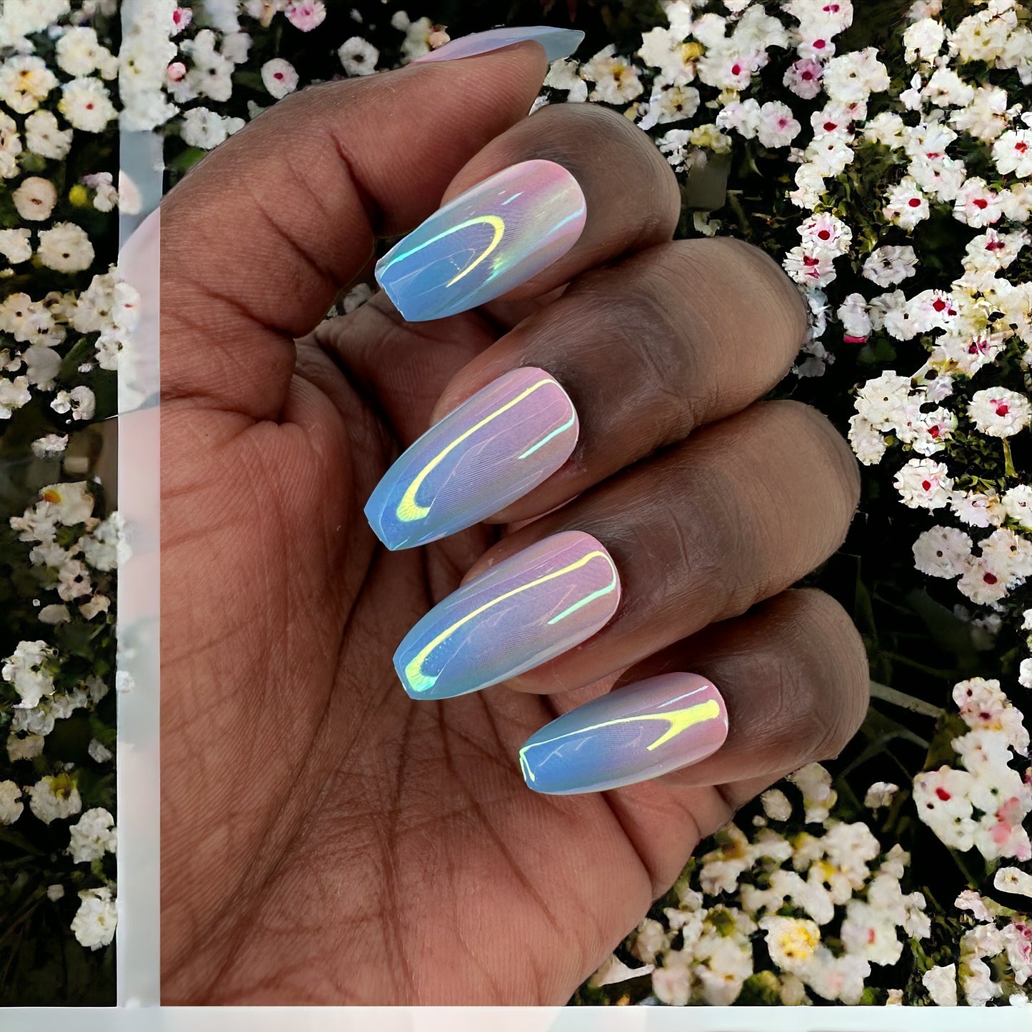 Fingers adorned with almond-shaped press-on nails in pink and blue chrome hues, set against a background of delicate white flowers